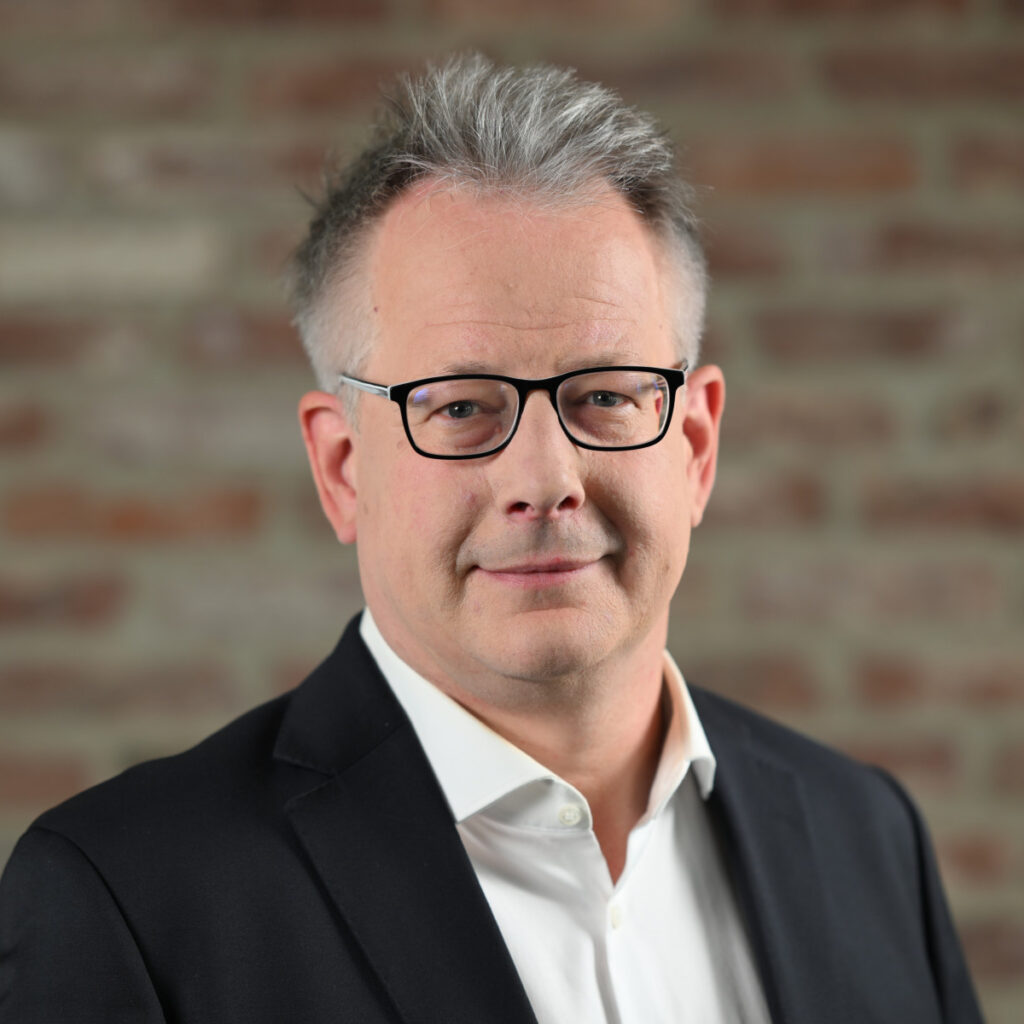 Porträtfoto von Alexander Kern, Chief Sales Officer. Er trägt eine schwarze Brille, einen weißen Hemdkragen und ein dunkles Sakko. Der Hintergrund ist eine unscharfe Ziegelsteinwand.