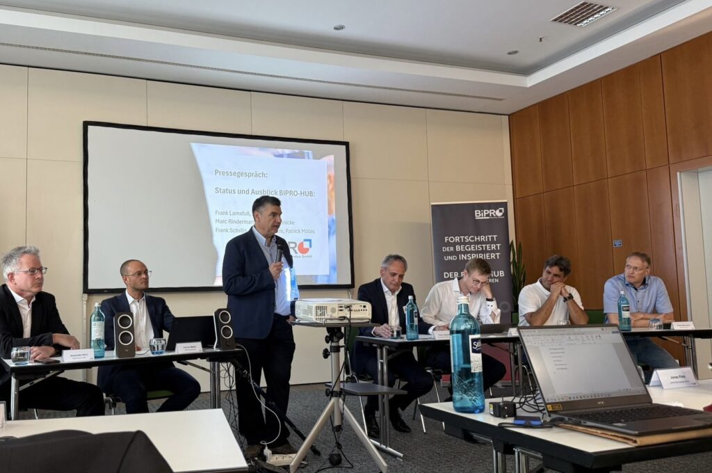 Foto einer Pressekonferenz in einem Konferenzraum. Sieben Personen sitzen an zwei Tischen, die zu einer U-Form angeordnet sind. Eine Person steht in der Mitte und spricht in ein Mikrofon. Im Hintergrund ist eine Leinwand mit der Projektion „Pressegespräch: Status und Ausblick BiPRO-HUB“ zu sehen. Auf einem Tisch steht ein Laptop, mehrere Wasserflaschen und Gläser. Ein Banner mit dem BiPRO-Logo und dem Text „Fortschritt der begeistert und inspiriert“ steht rechts im Raum.
