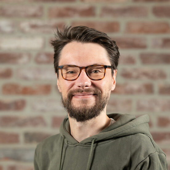 Porträt eines lächelnden Mannes (Florian Höser) mit Brille, braunem Vollbart und dunkelgrünem Hoodie vor einer Backsteinwand.